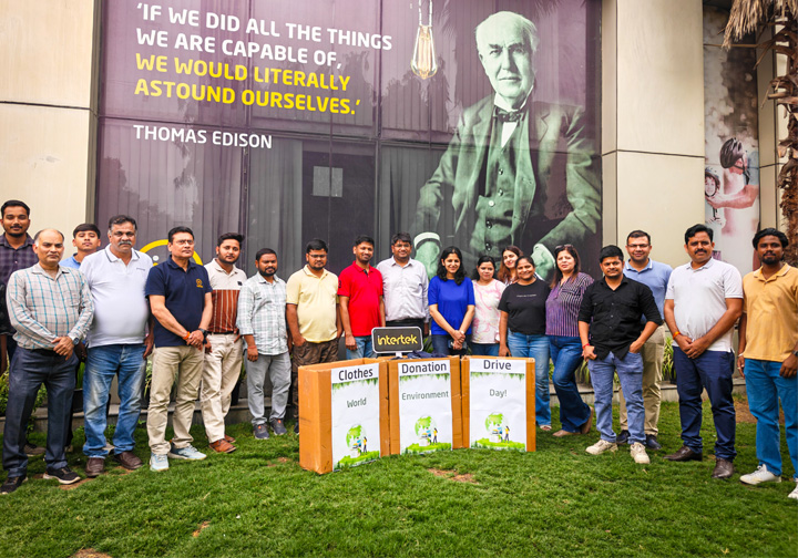 A large group of Intertek India colleagues stands behind marked clothes donation boxes. 