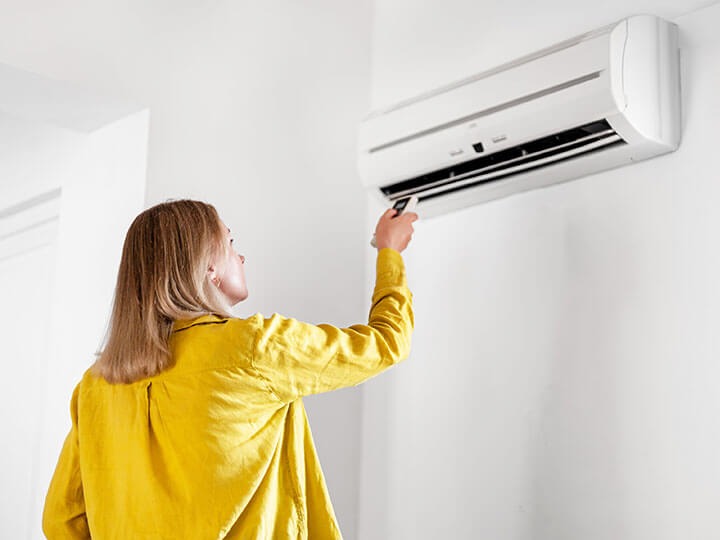 Rear view of woman in golden yellow long sleeve shirt aiming a remote at an air conditioning unit high on a wall near the ceiling.