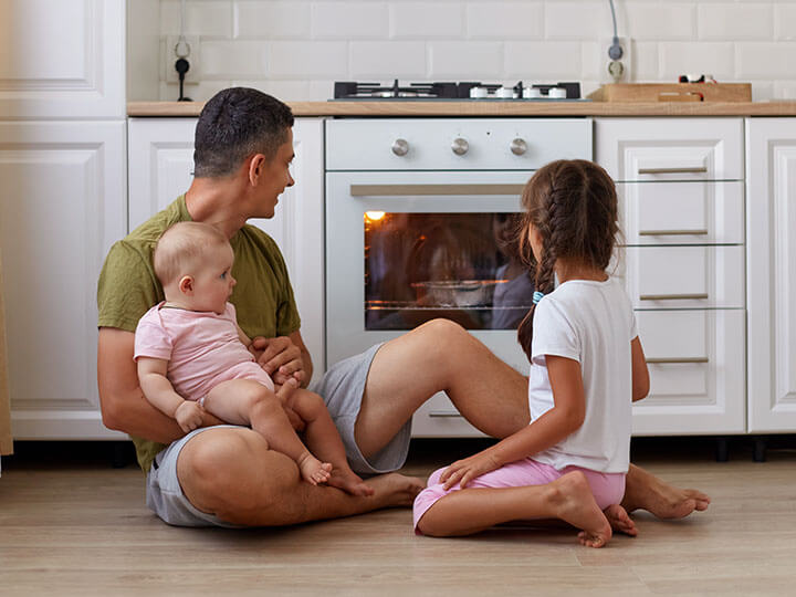 man wearing casual shirt and shorts sitting on the kitchen floor with his daughters watching food bake in the oven