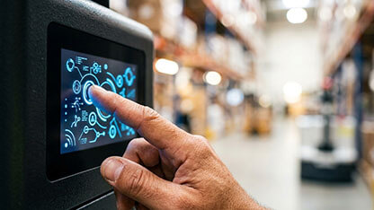 Close-up of a hand touching a digital control interface on a touchscreen panel displaying data visualizations and schematics inside a large industrial warehouse distribution center.